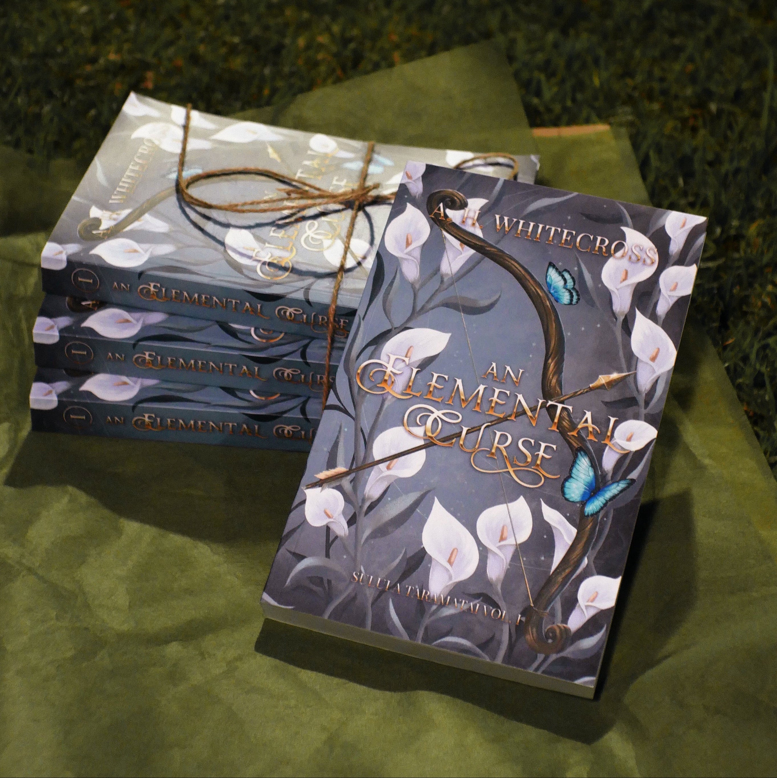 Books titled 'An Elemental Curse' by A. H. Whitecross on green paper and grass.